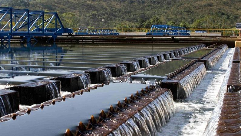 Estação de tratamento de água: fileiras de vertedouros com água cristalina caindo, estruturas azuis e paisagem montanhosa.