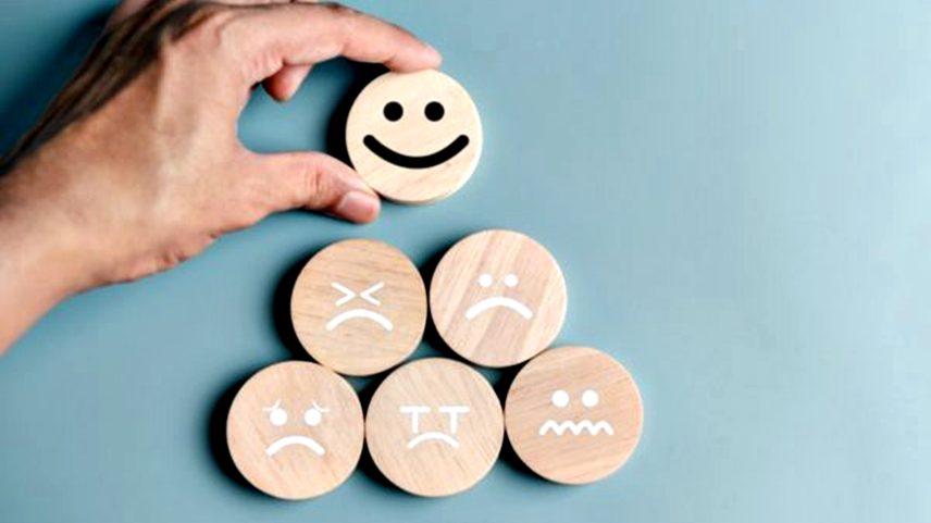 Mão de homem colocando uma carinha feliz de madeira próximo a outras carinhas de diferentes emoções de madeira formando uma pirâmide em fundo azul claro - Metrópoles
