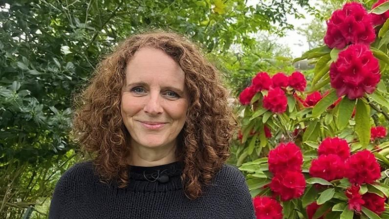 Maggie O'Farrell with curly auburn hair in black jumper. Background shows leaves on left and red flowers on the right