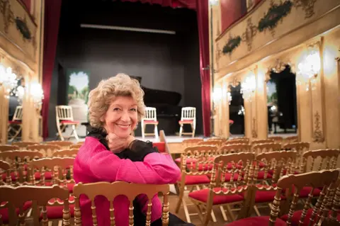 Getty A woman with short curly hair dressed in black and pink leans on the back of a chair and smiles to a camera while sitting in a theatre space.