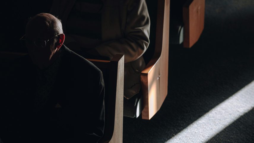 A close up image of people sitting in the pews of a dark church with a ray of sunshine.