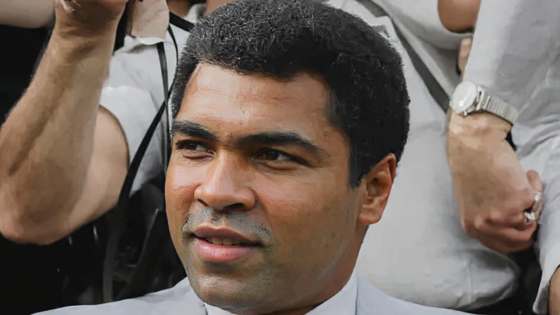 Richard Blanshard A black and white image of Muhammad Ali smiling and looking to the left of the camera. He is wearing a suit and behind him is a photographer holding a camera.