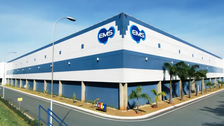 Fachada de um grande edifício industrial nas cores branco e azul com logos azuis da "EMS", palmeiras ao longo da calçada e céu azul ao fundo.