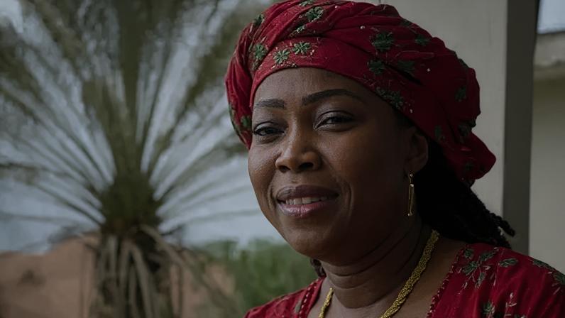 BBC Headshot of a smiling woman. She is wearing a red headwrap and matching top which are patterned with green flowers. She has a gold necklace and earrings.