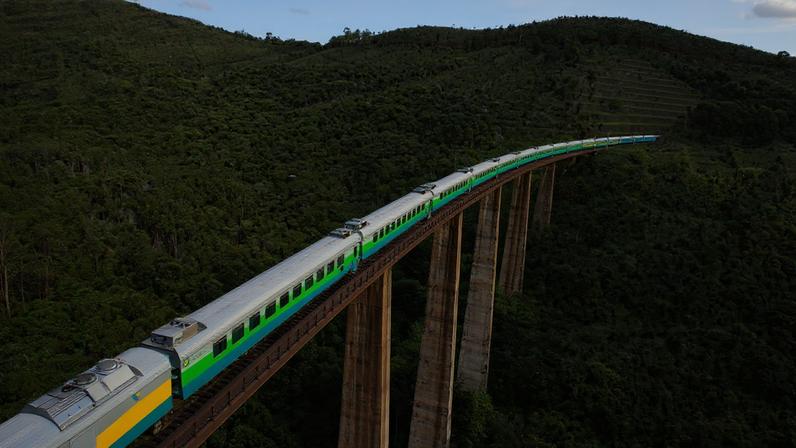 Trem de passageiros atravessa uma ponte ferroviária elevada sobre uma área de mata e morros verdes, em região montanhosa do Brasil. A composição faz uma curva em meio à paisagem rural.