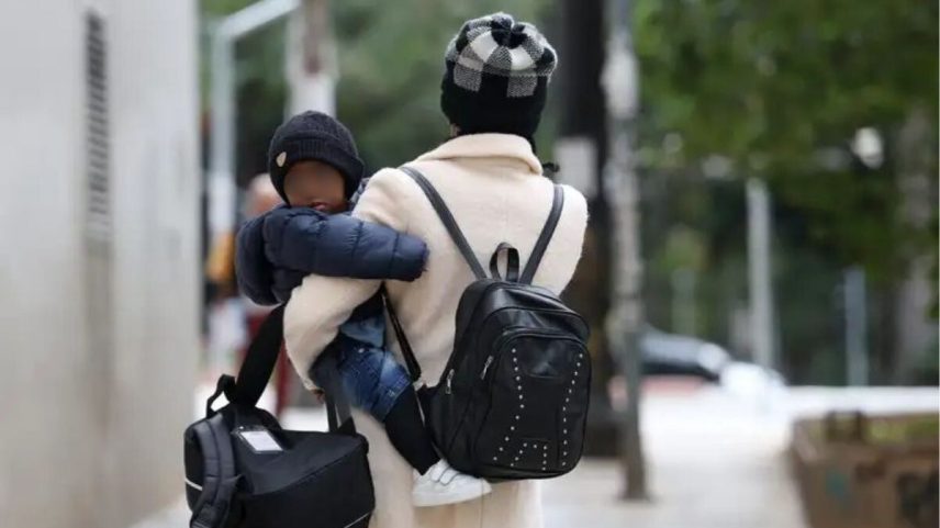 Imagem colorida mostra mulher de costas, caminhando. Ela veste casaco de frio e gorro na cabeça e carrega um bebê nos braços - Metrópoles