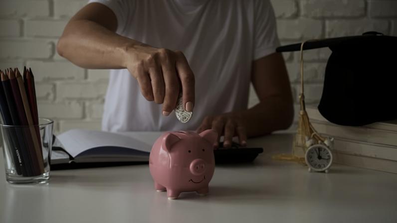 Pessoa coloca moeda em um porco roda de porcelana em cima de uma mesa, com caderno, lápis e relógio também em cima dela. A pessoa também digita em uma calculadora.