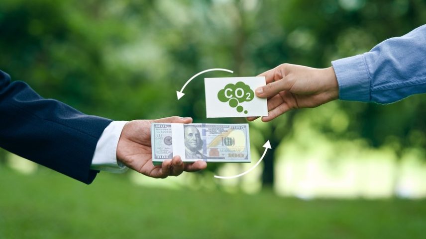 Pessoa segurando uma nota de dólar e outra segurando uma placa com o ícone de CO2, para ilustrar o conceito de créditos de carbono.