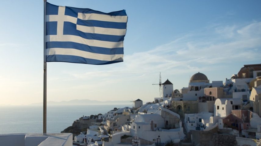 Imagem colorida da bandeira da Grécia, em destaque, tremulando em uma dia de céu azul. Ao fundo, o mar e um conjunto de casas em um cenário paradisíaco - Metrópoles