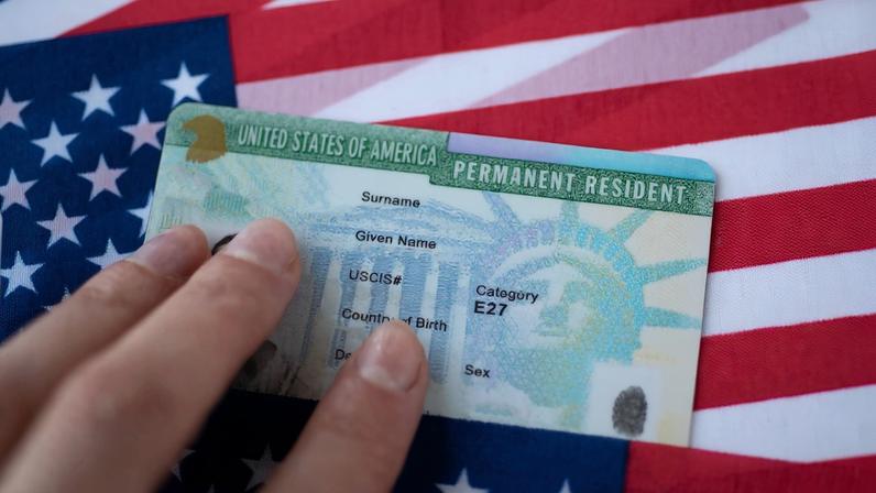 Foto colotrida de green card de residente permanente dos Estados Unidos, sobre a bandeira norte-americana - Metrópoles