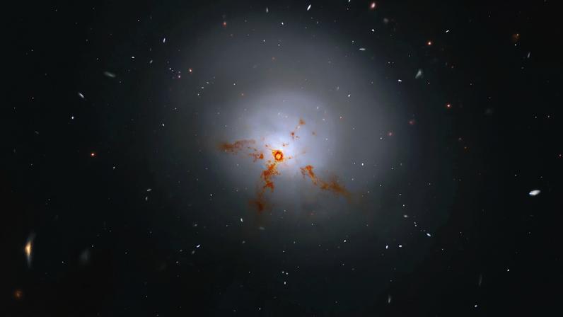 This Hubble image is of a bright lenticular galaxy (NGC 1266) seen nearly face on. Broad bright and dimmer areas of light hint at spiral structure, but there are no distinct spiral arms. Rusty-reddish-brown dust bisects the galaxy. More distant galaxies dot the black background, some even shining through the less dense regions of the bright foreground lenticular galaxy.