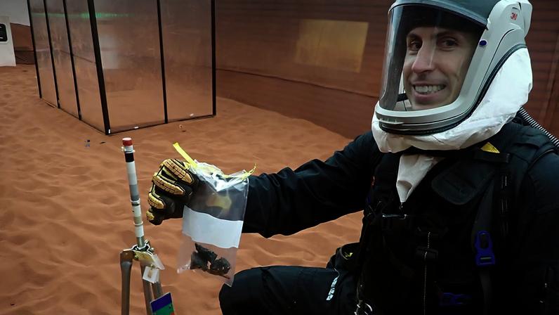 HWHAP Ep. 423. A member of the CHAPEA 2 crew wearing a simulated space suit poses next to a small structure on the simulated surface of Mars.