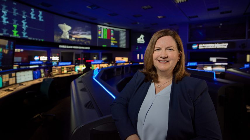 Woman stands in a mission control room, with rows of computer consoles and large overhead screens displaying antenna imagery, data charts, the Artemis logo, and communications systems in operation. The Jet Propulsion Laboratory placard is visible in the background.