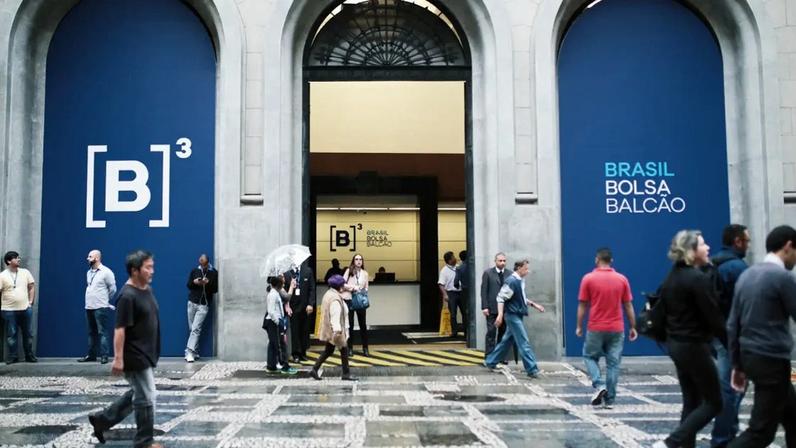 A imagem mostra a movimentada entrada da B3, a bolsa de valores brasileira, com fachada clássica e painéis laterais azuis destacando o logotipo [B]³ e o texto "BRASIL BOLSA BALCÃO", enquanto várias pessoas transitam em trajes casuais e de trabalho.