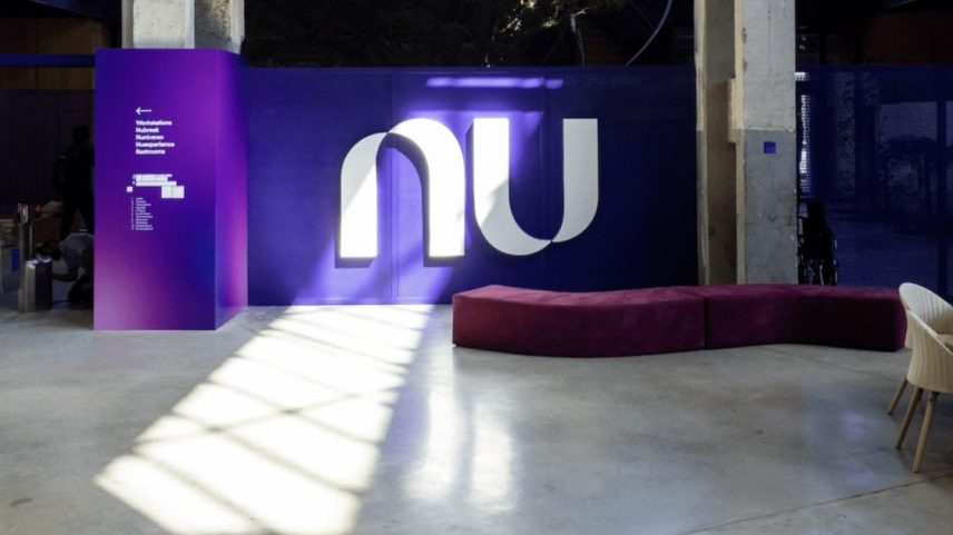 Interior de escritório roxo com logo 'nu', banco e sinalização direcional. Sombra no chão de concreto e pessoa perto de catraca.