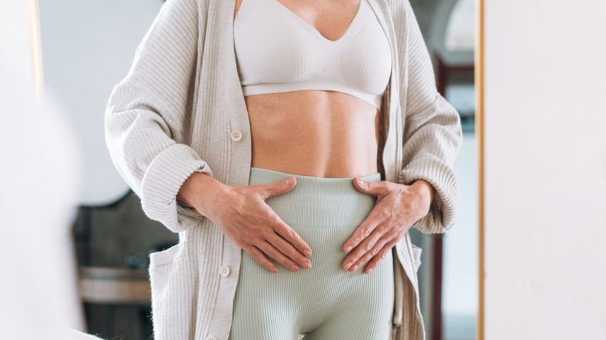 Foto colorida de mulher se olhando no espelho e com a mão na barriga - Metrópoles