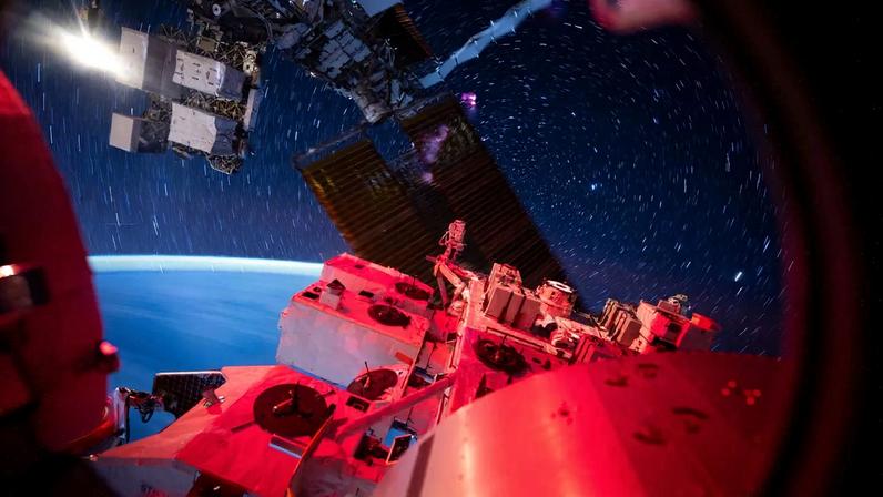 View from the International Space Station showing its illuminated exterior hardware in red light against a backdrop of deep space. Star trails form circular streaks across the sky due to the station’s motion, while Earth’s blue curvature glows softly at the horizon.