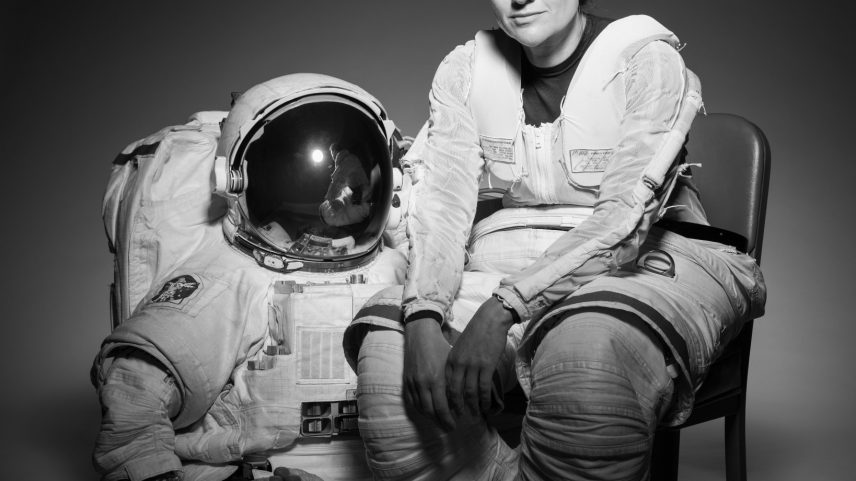 NASA Astronaut Jessica Meir sits in a chair. Her body is slightly turned to the right as she looks forward. She is wearing the bottom half of a spacesuit; the top half and gloves are on the floor next to the chair.