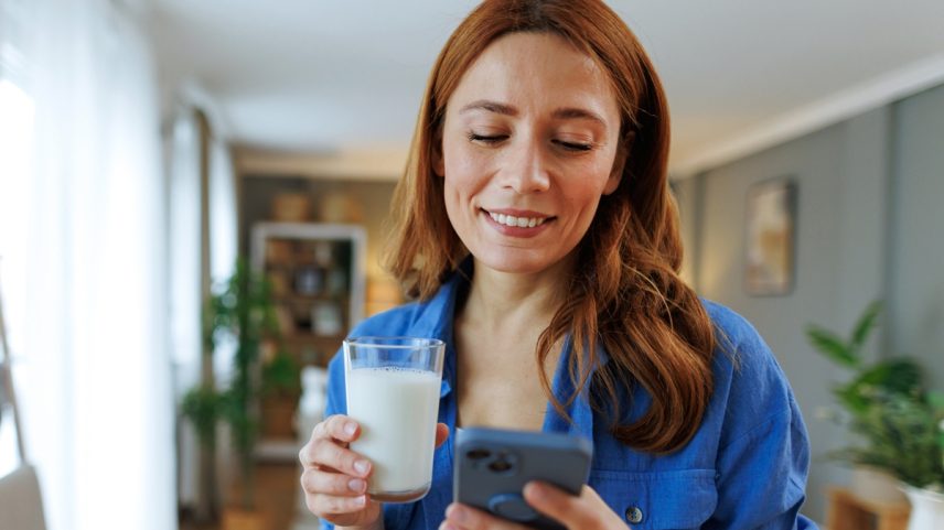 Foto colorida de mulher com blusa azul segurando um copo de leite e teclando no celular - Metrópoles