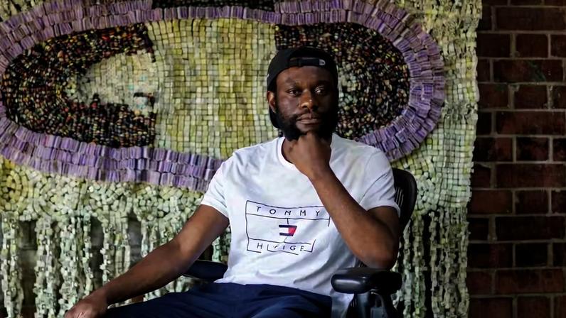 O artista zimbabuano Moffat Takadiwa posa diante de uma de suas tapeçarias de resíduos plásticos; obras inéditas estão em exposição na Galeria Almeida & Dale, em São Paulo