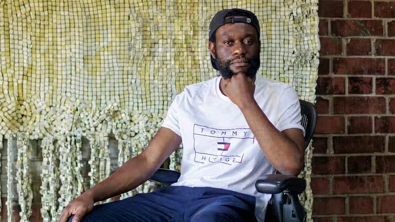 O artista zimbabuano Moffat Takadiwa posa diante de uma de suas tapeçarias de resíduos plásticos; obras inéditas estão em exposição na Galeria Almeida & Dale, em São Paulo