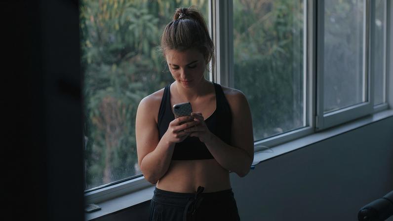 Foto mostra mulher fitness, na academia, mexendo no celular. Vendo postagens fitness. Metrópoles