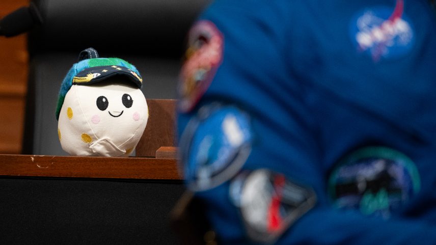 Rise, the Artemis II zero gravity indicator, sits on a wooden desk. A NASA astronaut is in the foreground.