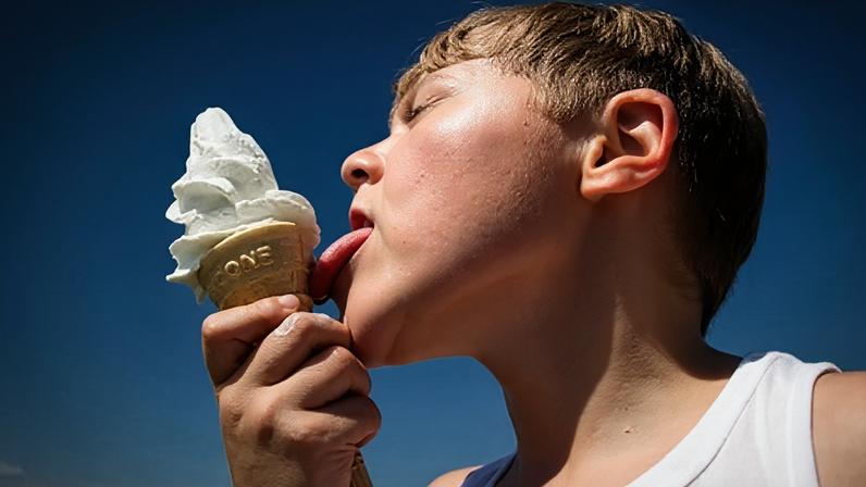 Young young boy in a white vest licks a soft srve vanilla icecream against the backdrop of a bright blue sky. (Credit: Getty Images)