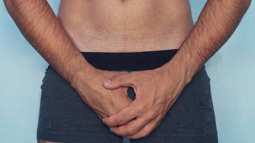 Foto colorida de um homem da cintura para baixo de cueca e cobrindo o pênis em um fundo azul - Metrópoles