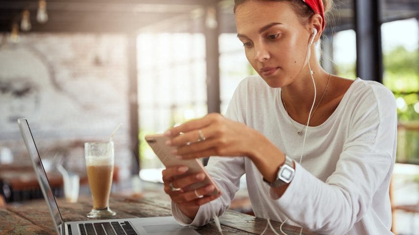 Mulher sentada em um cafeteria com fones de ouvidos mexendo no celular. Metrópoles