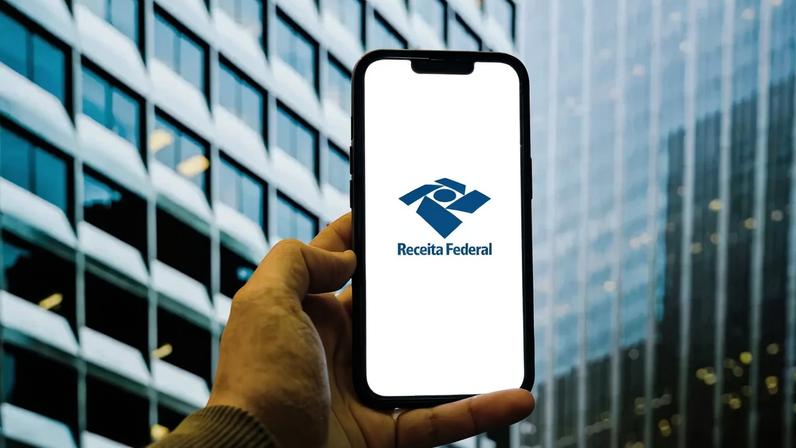 Mão segura smartphone com app da Receita Federal, em frente a um prédio moderno.