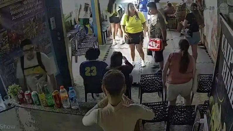 Pessoas em um evento de capoeira. Um homem de óculos e mochila clara está atrás de um balcão com bebidas. Outras pessoas estão sentadas em cadeiras ou em pé, conversando. Uma mulher de camiseta amarela e saia preta está no centro, enquanto outra de camiseta vermelha e shorts claros está de costas. O ambiente parece movimentado e informal