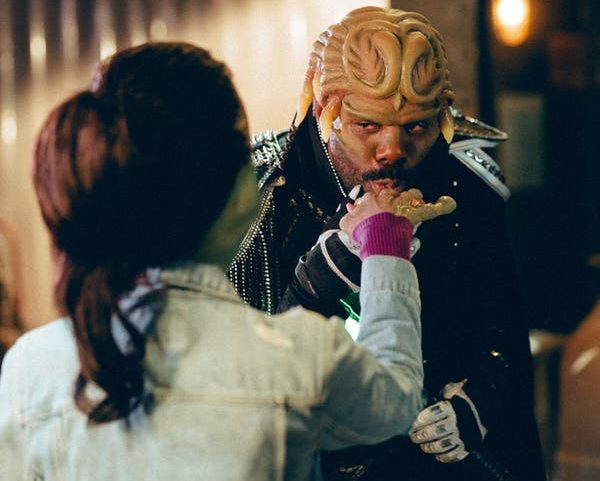 A man costumed as an alien warlord with a lizard-like head kisses the very large, frog-like hand of a woman whose face we can't see.