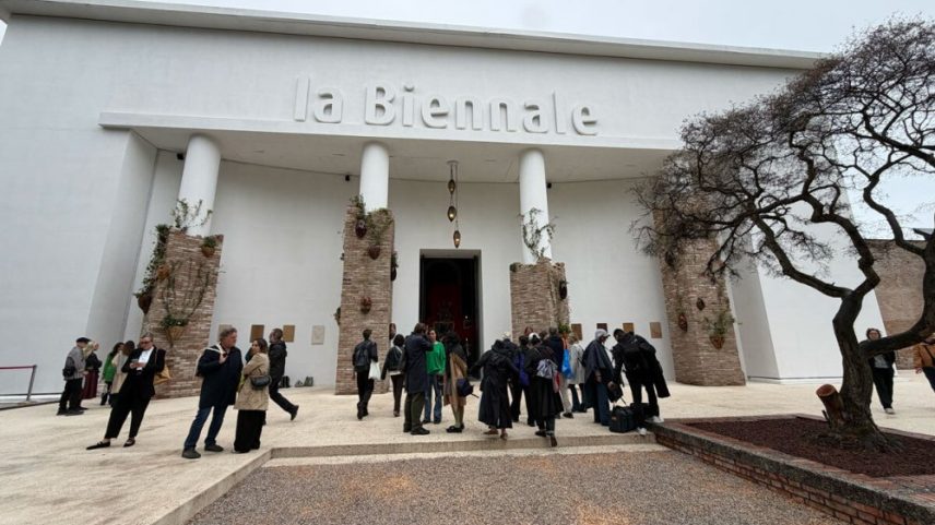People stand in front of a white structure that reads 'la Biennale'; its four columns have been covered with bricks and flowering plants.
