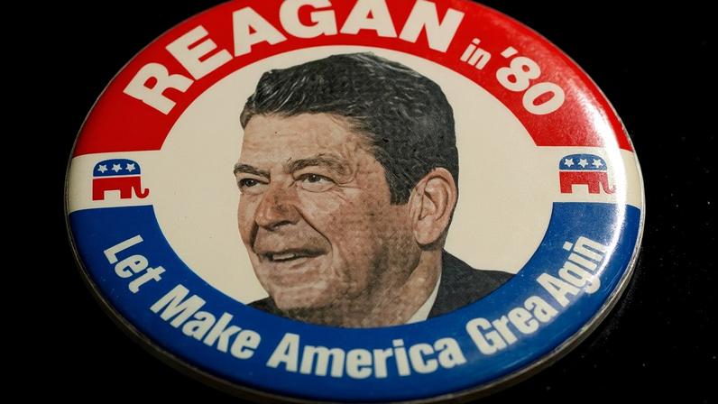 A campaign button for Ronald Reagan. In white text on a red background, it says “Reagan in ‘80” and then in the bottom of the button, it says, “Let’s Make America Great Again” in white text on a blue background. A black and white photo of Ronald Reagan’s face and shoulders in a suit is in the middle of the pin.