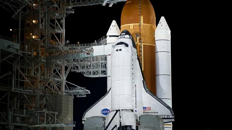 Ônibus espacial Endeavour branco acoplado a um tanque de combustível laranja e dois propulsores brancos, em uma plataforma de lançamento iluminada à noite