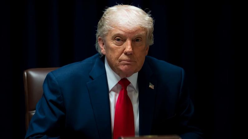 President Trump sitting during a meeting. A binder is open on the table in front of him.
