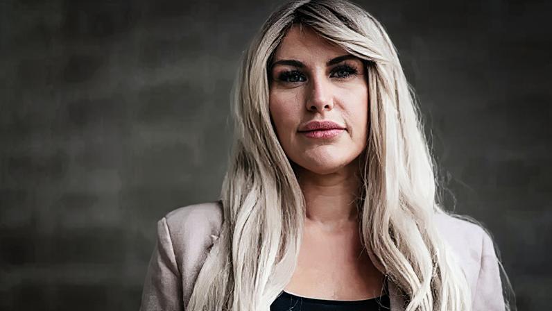 Gus Palmer/BBC A head and shoulders portrait of Rebecca, standing against a light stone background. She has long blonde wavey hair and wears a black top with a light pink jacket. She looks pensively into the camera.