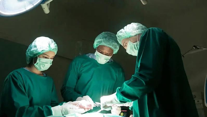 Três cirurgiões, dois homens e uma mulher, usando máscaras e toucas cirúrgicas verdes, operam sob luzes fortes em um centro cirúrgico. A mulher à esquerda segura um tecido, enquanto o homem à direita usa pinças. Um monitor exibe "80" em destaque
