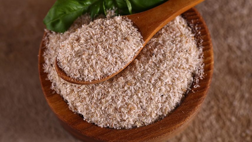 Fibras de psyllium em uma tigela de madeira com uma colher de pau cheia, e folhas verdes de manjericão ao fundo