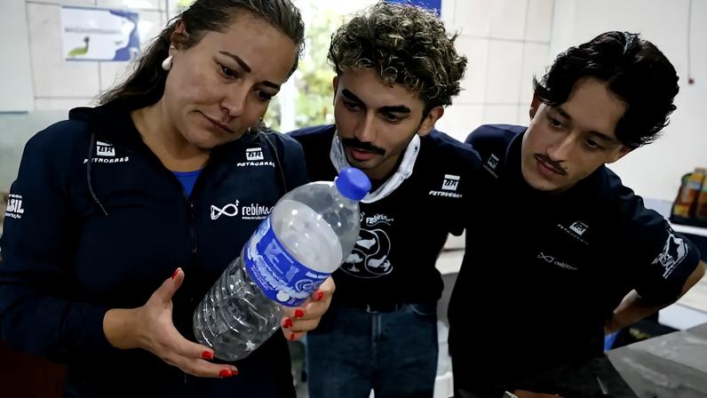 Mulher de blusa azul escura com logo Petrobras e Rebinma, unhas vermelhas, segura uma garrafa plástica transparente com líquido branco. Dois homens jovens, um com cabelo cacheado e outro com bigode, ambos de camisetas escuras com logos Petrobras e Rebinma, observam a garrafa atentamente