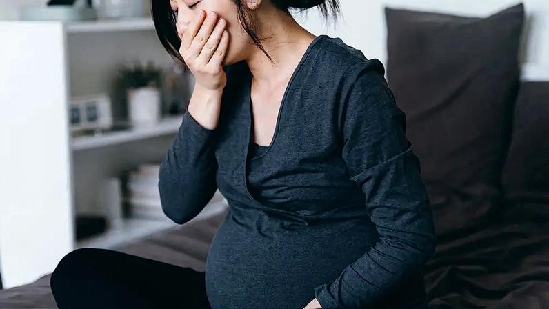 Mulher grávida asiática, de blusa cinza e calça preta, sentada na cama, com a mão na boca, parecendo enjoada ou com náuseas