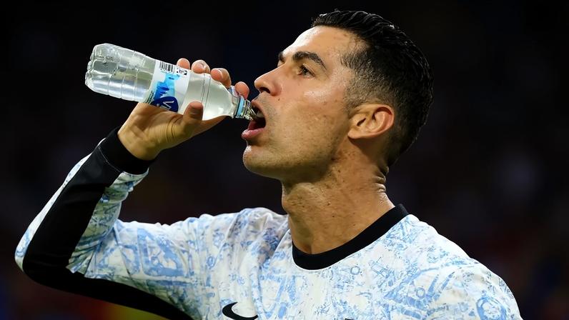 Cristiano Ronaldo, suado, bebe água de uma garrafa plástica transparente, vestindo camisa de manga comprida azul e branca com detalhes pretos, em um estádio lotado