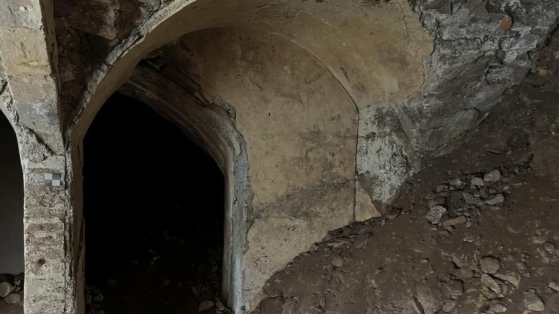 Entrada de um túnel escuro com arco de pedra e argamassa desgastada, cercado por terra e detritos. Na parede direita, grafite vermelho INDIANA JONES 2013