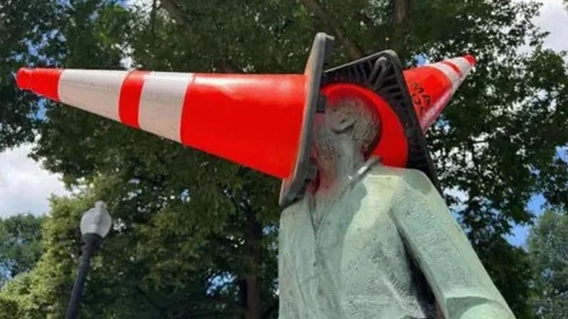 Alamy A person in a park wearing a blue hat and a sleeveless sports jersey with the number 23 places a bright orange traffic cone on top of a seated statue’s head, which is already covered by a larger cone. The statue sits on a decorative base surrounded by grass and trees, with city buildings visible in the background on a sunny day.
