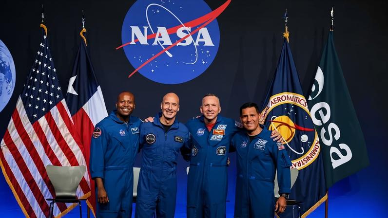 Quatro astronautas sorridentes, vestindo macacões azuis, posam abraçados em frente a bandeiras dos EUA, Texas, NASA e ESA