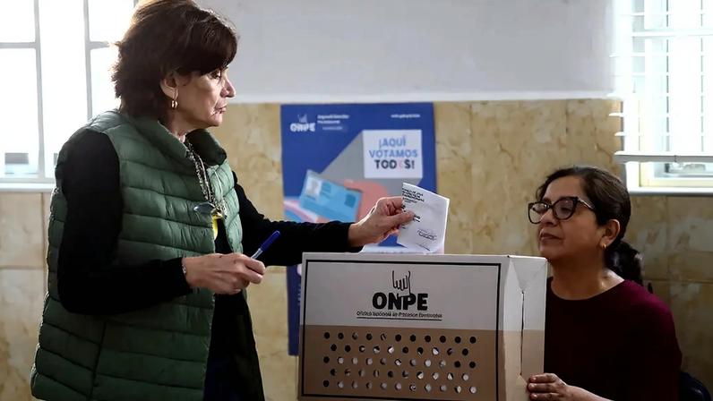 Mulher de colete verde escuro e blusa preta depositando um voto em uma urna eleitoral de papelão, enquanto outra mulher de óculos e blusa vinho observa. A urna tem o logo da ONPE e um cartaz azul ao fundo diz ¡Aquí votamos todos!.