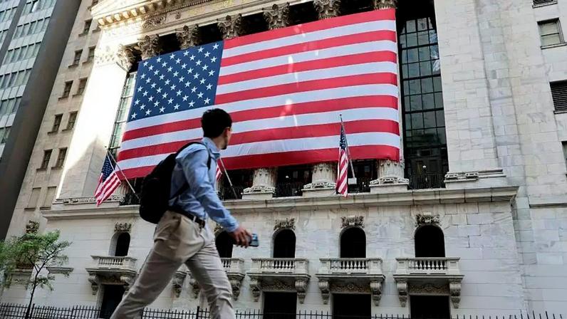A Bolsa de Valores de Nova York, nos Estados Unidos, em manhã de negócios no dia 26 de maio de 2026, com uma grande bandeira americana na frente do edifício e pessoas andando na calçada