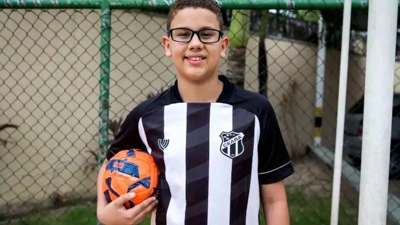 Um menino usando óculos de grau e uma camisa de futebol do Ceará. Ele segura uma bola de futebol com a mão direita.