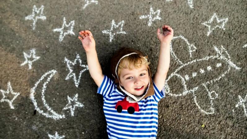 Menino sorridente usando chapéu de piloto e óculos de proteção, deitada no asfalto pintado com desenhos de avião, lua e estrelas.